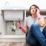 Woman with pink rubber gloves fixing plumbing under kitchen sink with pipe wrench, part of a home plumbing repair.