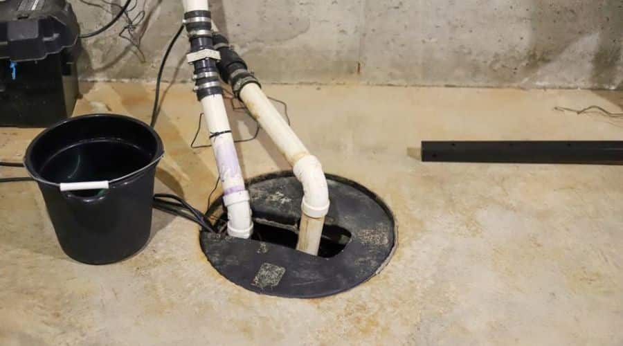 Sump pump setup with PVC pipes and a bucket in a concrete basement, highlighting plumbing service for water management.